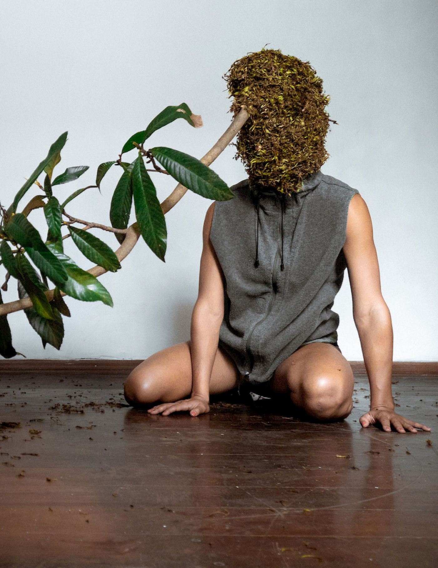 Kneeling person has a bonnet of moss pulled over the head, a branch with leaves seems to be growing out of the head.