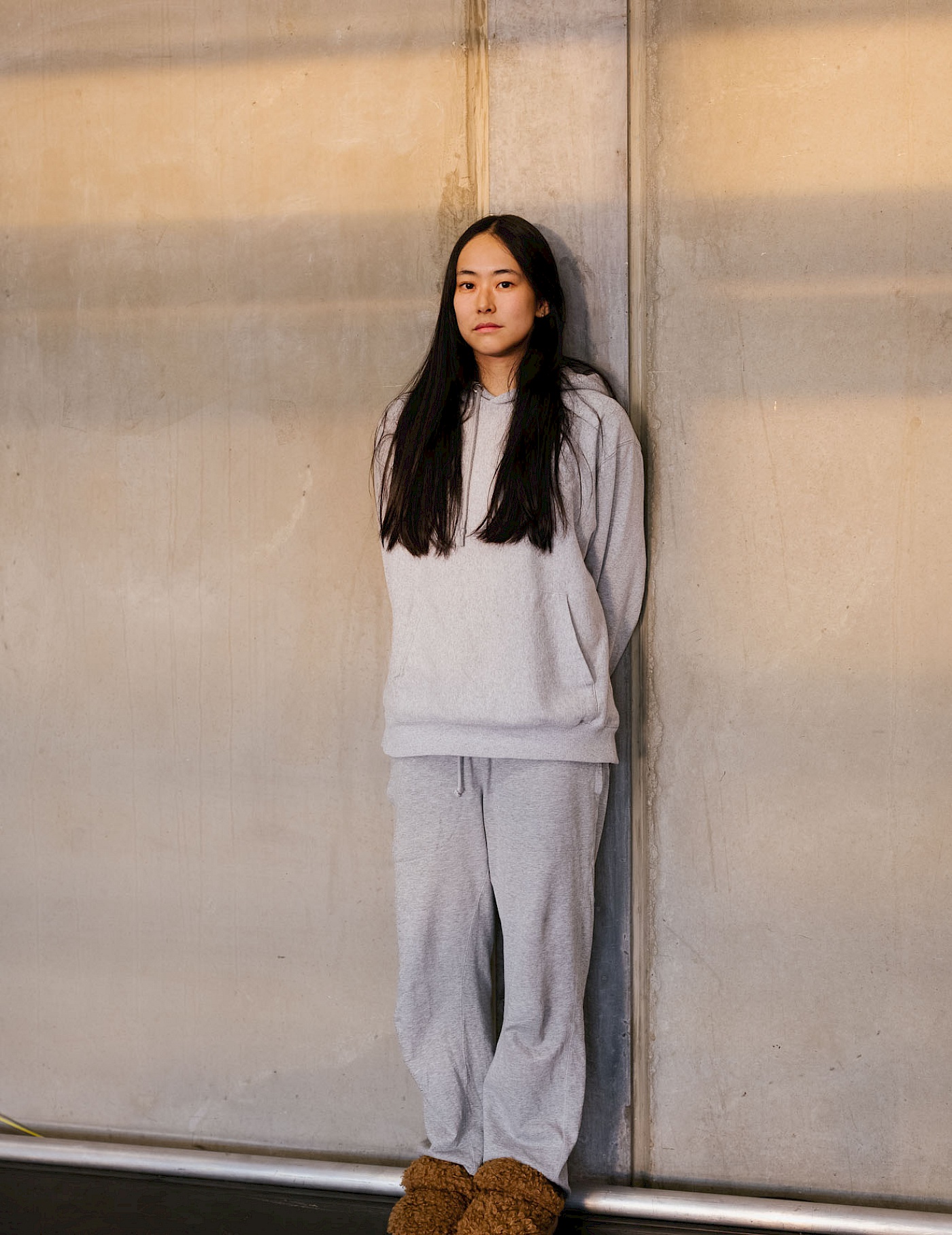 Phil Dera Hanako Hayakawa in grey jogging bottoms and a grey jumper in front of a grey concrete wall.