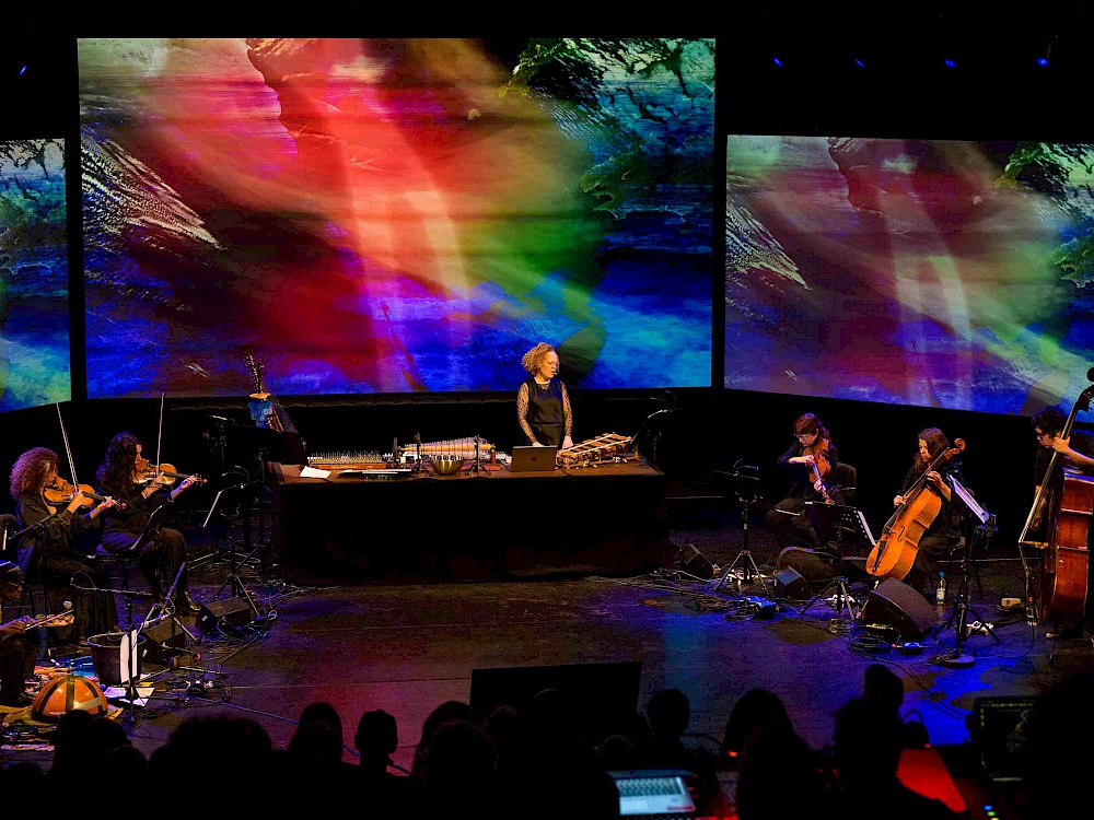 Bühnenansicht: Mehrere Musiker*innen im Kreis, dahinter eine farbenfrohe Videoprojektion.