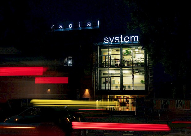 Nachansicht des Radialsystems mit Leuchtschrift und verwischten Autoscheinwerfern im Vordergrund