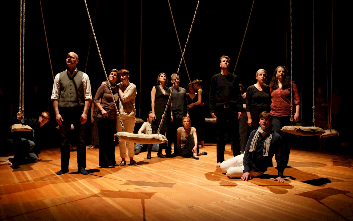 Matthias Heyde Eine Gruppe von Menschen steht und sitzt auf einer Bühne mit hängenden Schaukeln, sie blicken ernsthaft und konzentriert in verschiedene Richtungen.