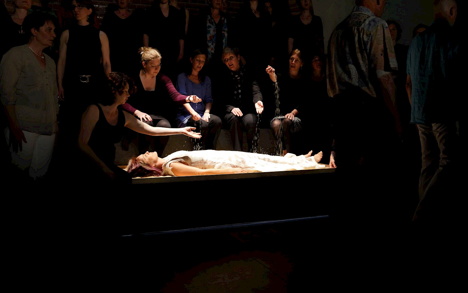 Matthias Heyde Eine Frau liegt in einem hellen Kleid aufgebahrt, während mehrere Menschen im Halbdunkel um sie herum versammelt sind.