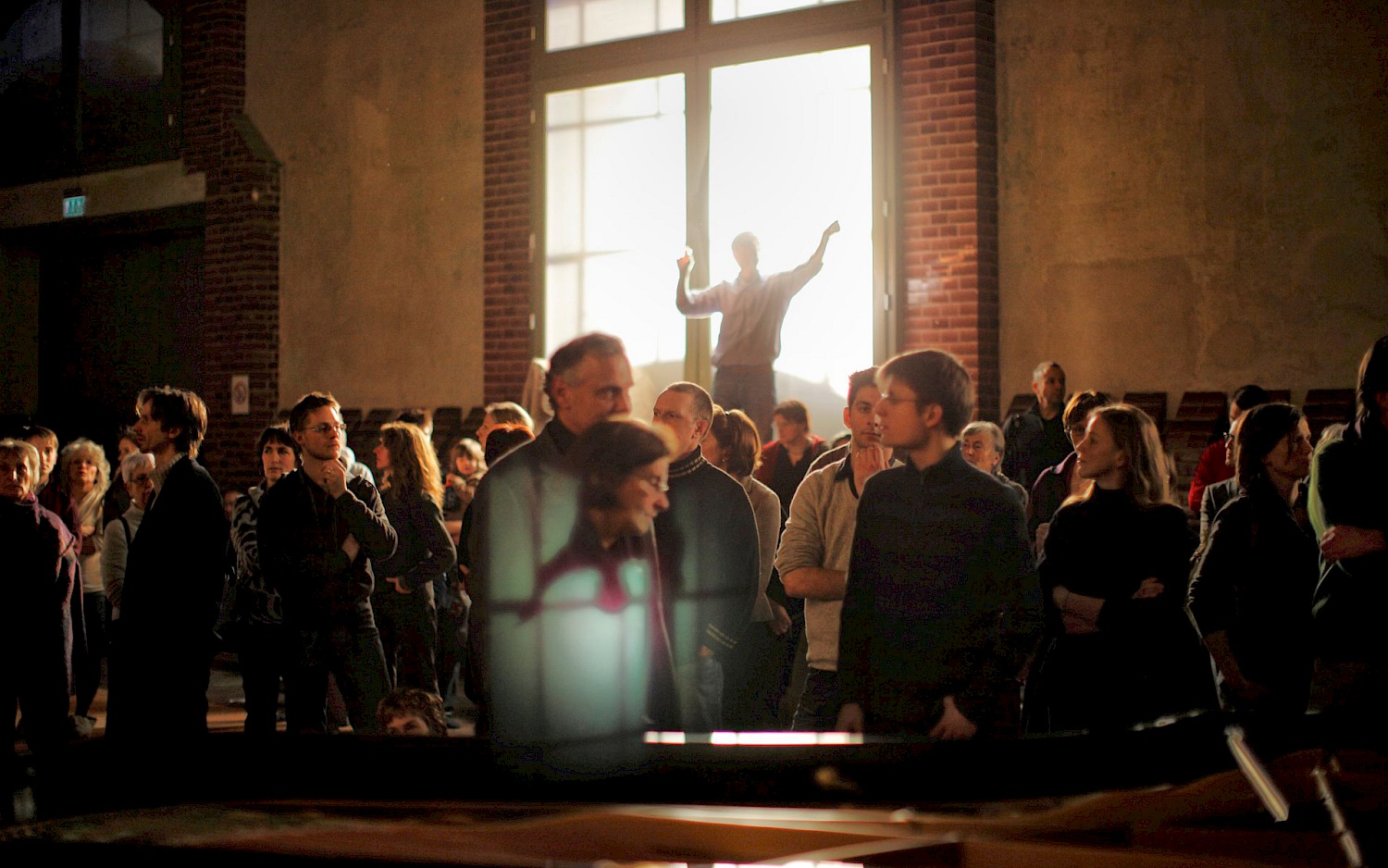 Matthias Heyde Über einer Ansammlung von Personen steht eine einzelne Person erhöht im Gegenlicht eines hellen Fensters und breitet die Arme aus.