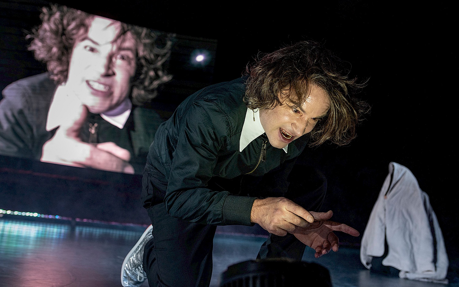Falk Wenzel Performer kniet auf der Bühne, im Hintergrund ein Live-Video von seinem drohenden Gesicht und ausgestreckter Finger