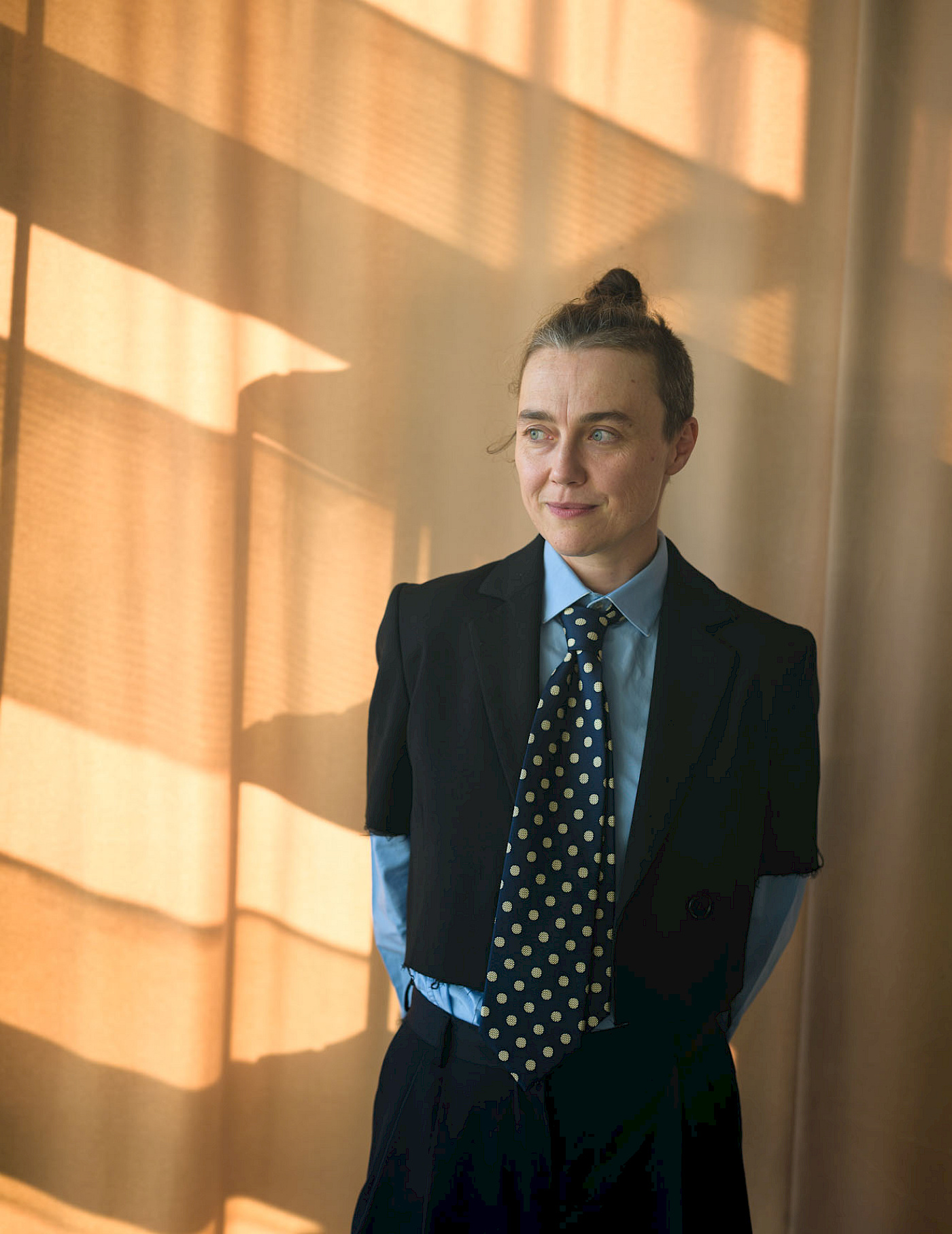 Phil Dera Agata Siniarska in schwarzem Blazer, blauem Hemd und gepunkteter Krawatte vor einem gelben Vorhang.