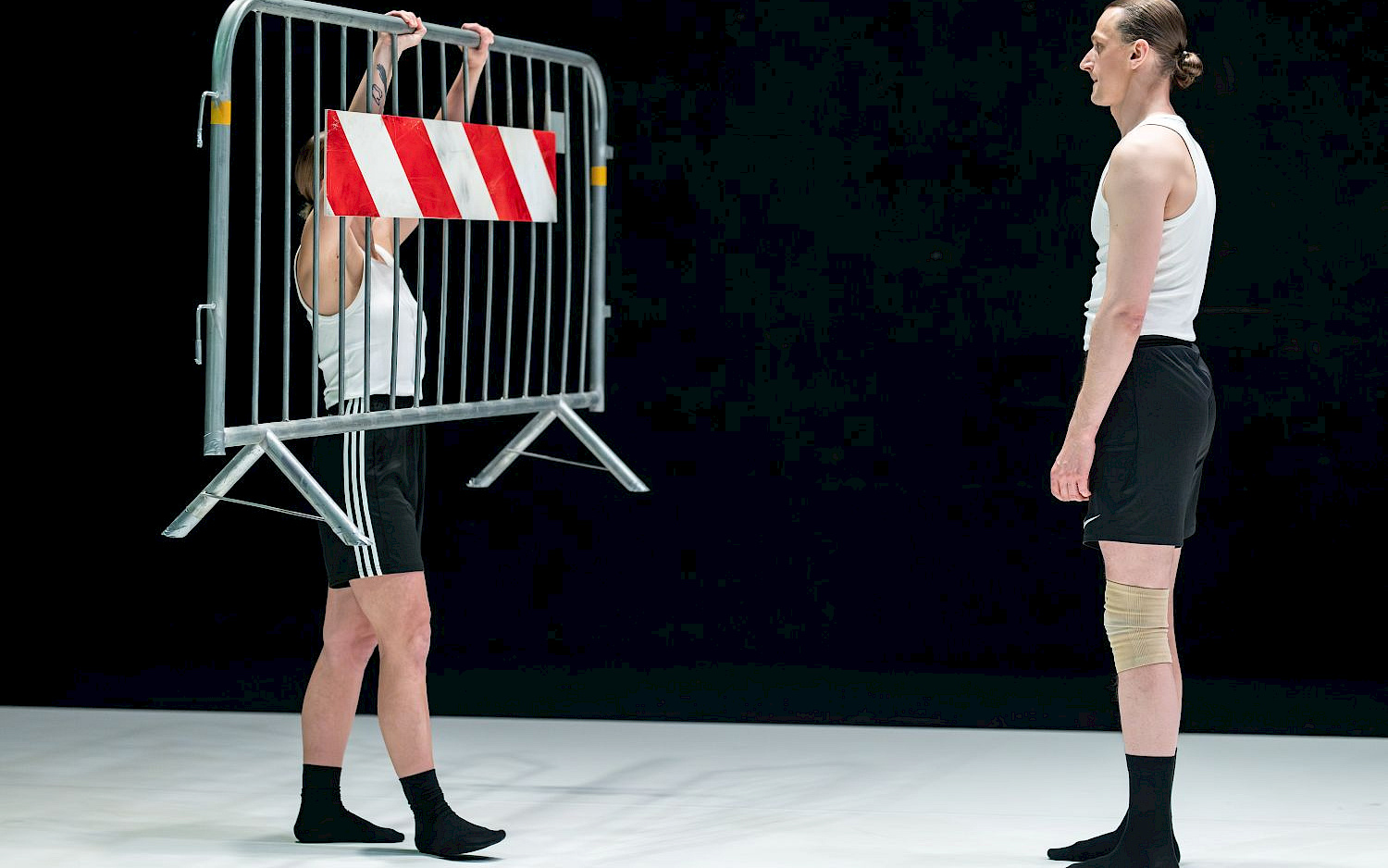 D. Alisauskas Zwei Performer*innen vor schwarzem Hintergrund, eine hält einen Absperrzaun in die Höhe.
