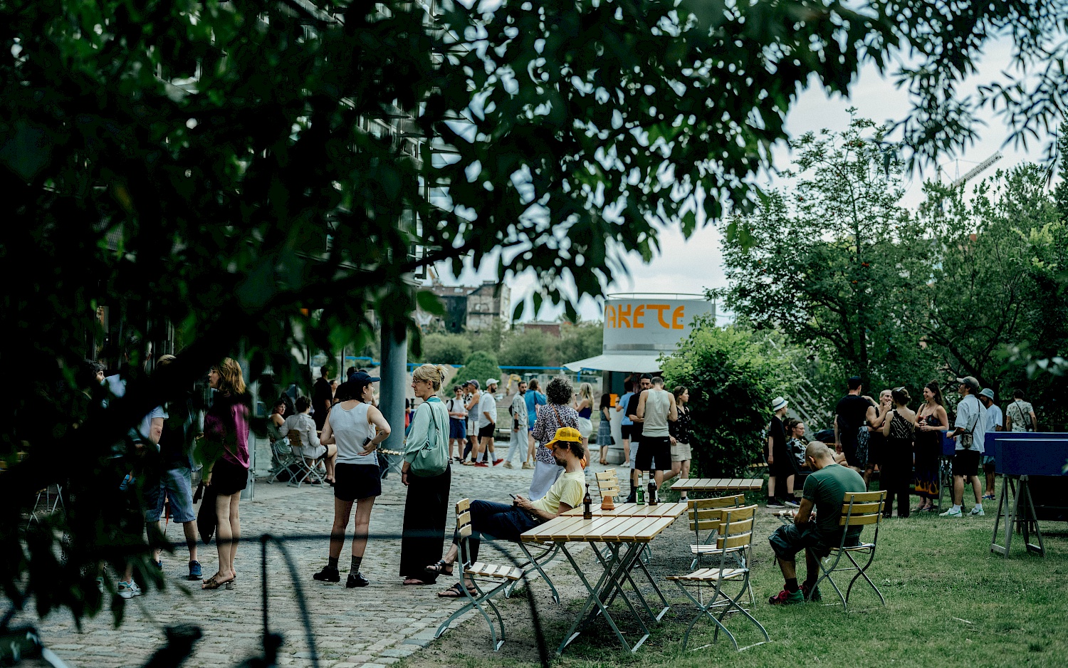 Sommerlich gekleidete Menschen stehen und sitzen im Garten und auf der Terrasse vom Radialsystem.