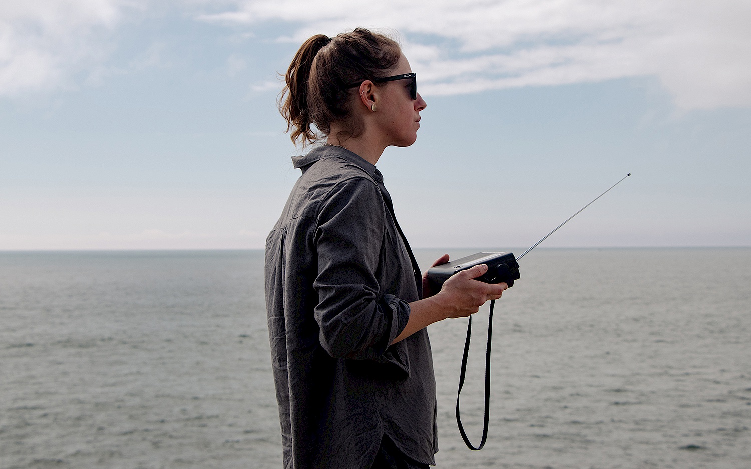 Florencia Curci steht seitlich vor unbewegtem Wasser und blauem Himmel. In der Hand hält sie ein kleines Radio.