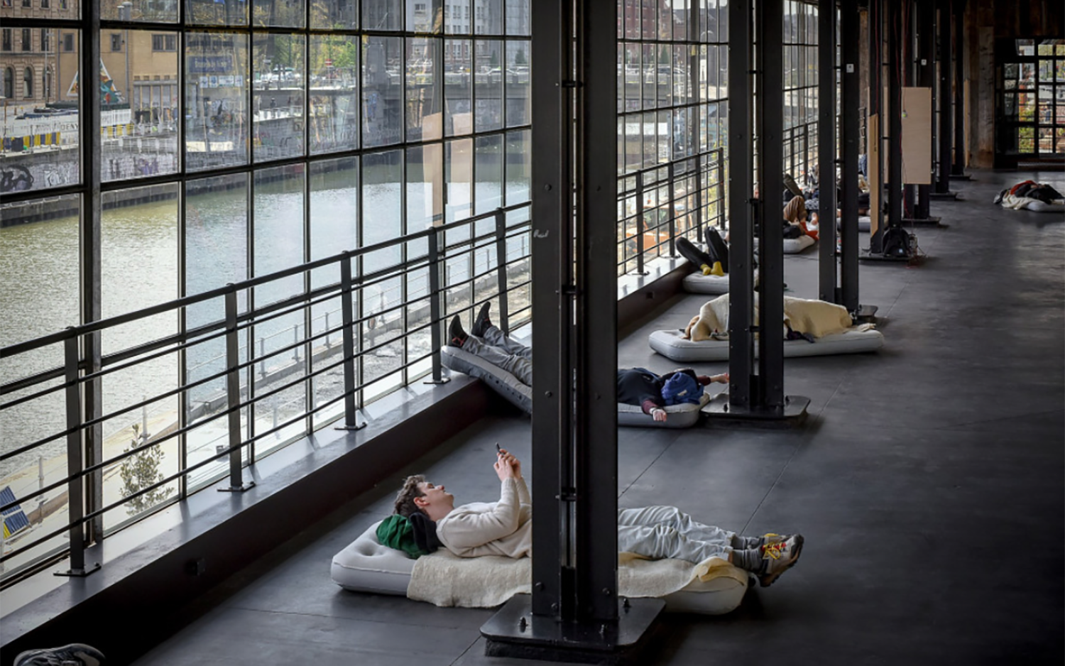 Mehrere Menschen liegen auf Luftmatratzen mit Decken und Kissen und besuchen die Performance Noctures for a Society in Brüssel.