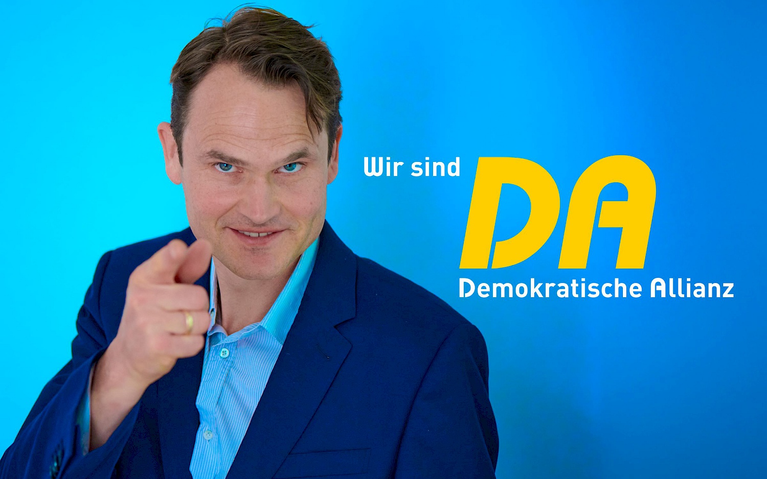 Ein Mann in blauem Anzug zeigt lächelnd mit dem Zeigefinger in die Kamera. Daneben vor blauem Hintergrund der Schriftzug: "Wir sind DA. Demokratische Allianz"